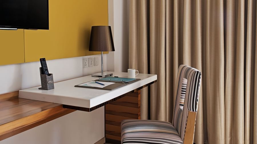 A corner view of the deluxe room at Barsana Boutique Hotel, Kolkata, showing a work desk, chair, wall-mounted television, and curtains.