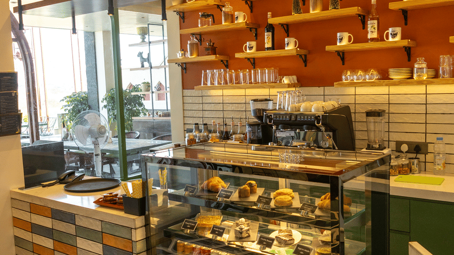 Cafe ordering section at Hotel Bawa, Udaipur showing a patterned front counter, glass food display case, coffee machine, and wall shelves.