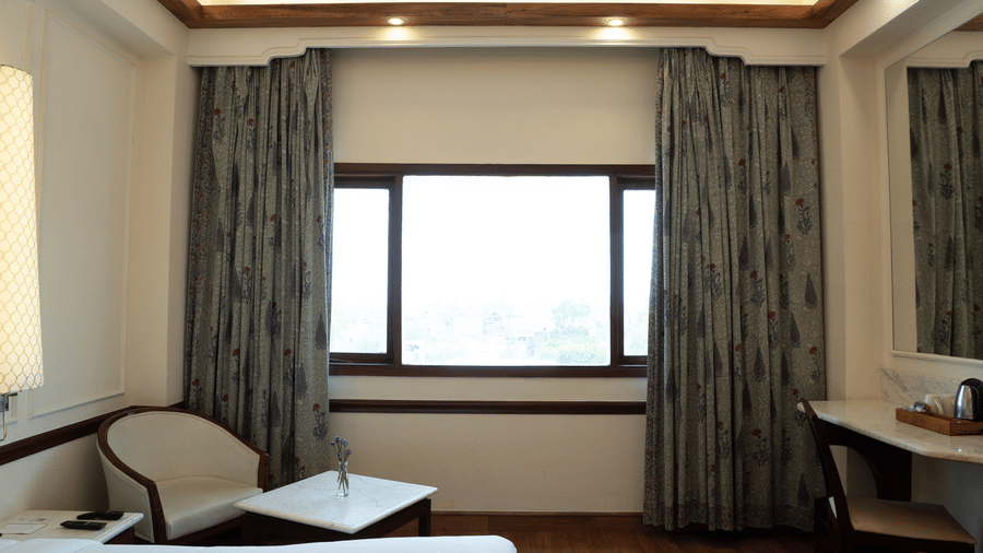 Hardwood floors lead toward a window and a desk area illuminated by recessed ceiling lights in the Deluxe King room at Clarks Amer, Jaipur.