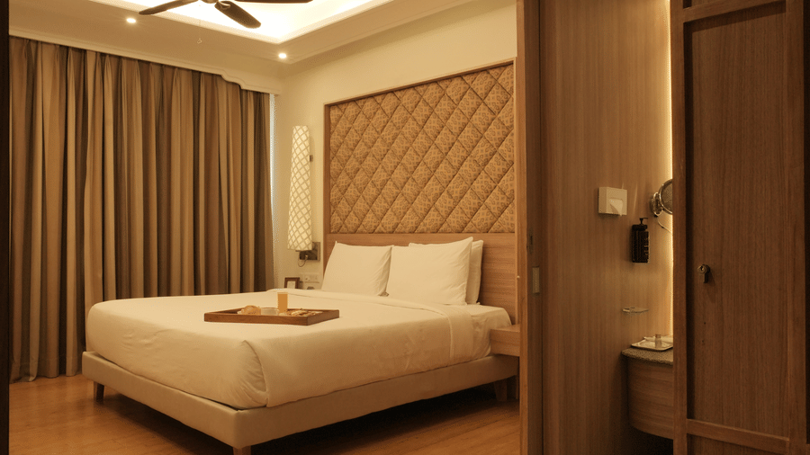 A bedroom with a king size bed, bedside tables, soft lighting, and curtains, viewed through a doorway in the Executive Suite at Clarks Amer, Jaipur.