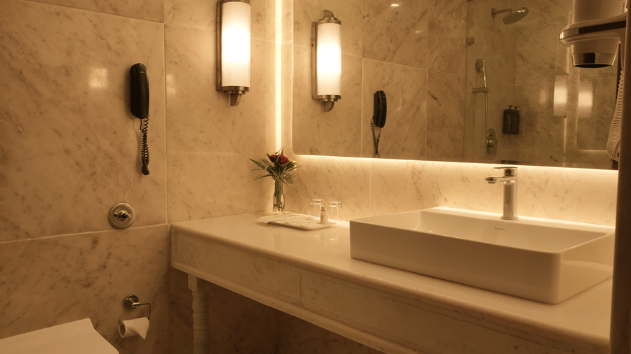 The bathroom in the Superior King at Clarks Amer, Jaipur features a long vanity with a rectangular sink, a large back-lit mirror, and stone walls.