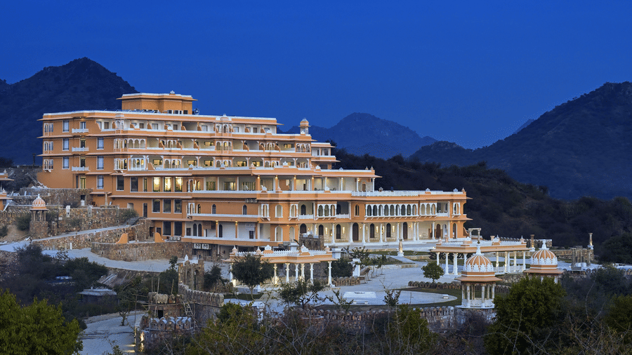 Facade of Fateh Vilas during twilight with numerous trees in the foreground and mountains in the background, ideal for a destination wedding in Udaipur