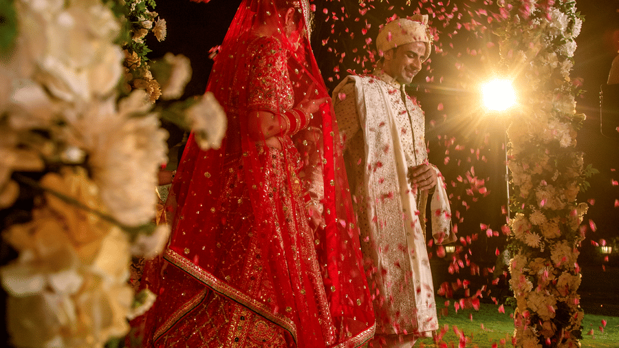 A bride in a red lehenga stands under a beautifully decorated floral canopy, her face covered with a traditional veil, surrounded by lush flowers and soft lighting.