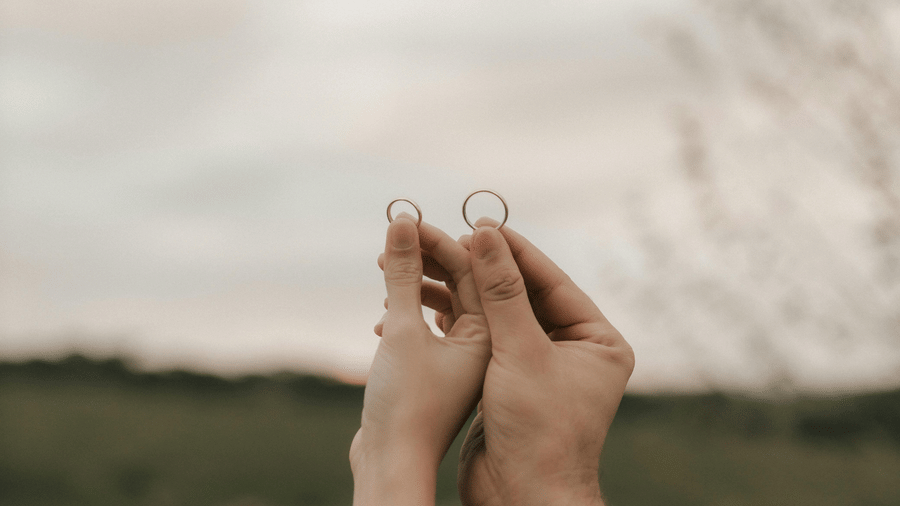 A close-up shot of 2 people holding hands in a field with a cloudy sky above.