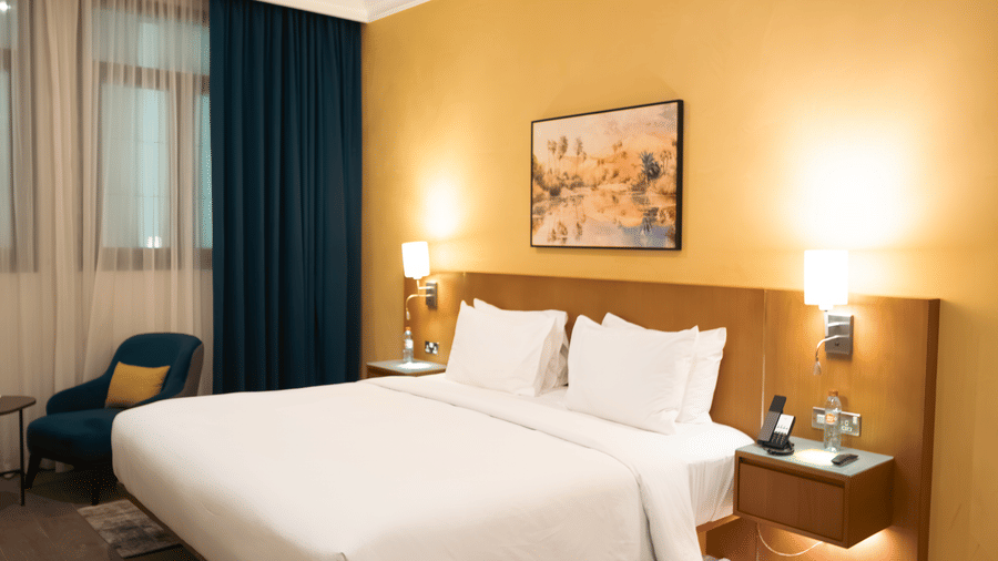 Large bed with white linens on a patterned rug, yellow accent wall, wood headboard, and a dark blue armchair near the window.