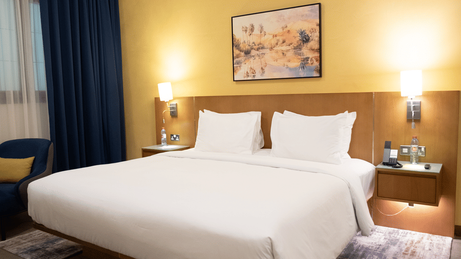 Large bed with white linens on a patterned rug, yellow accent wall, wood headboard, and a dark blue armchair near the window.
