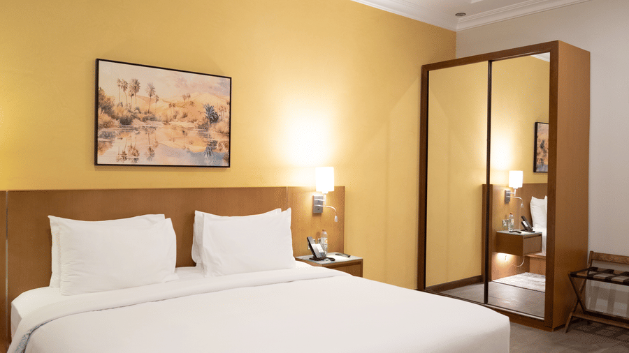 Bedroom with yellow wall, white bed, wood headboard, and a large mirrored wardrobe reflecting the room. Dark wood flooring.