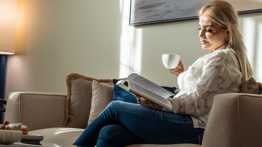 Woman with blonde hair reading a book and sipping coffee on a beige sofa in a sunlit living room with a large desert painting.