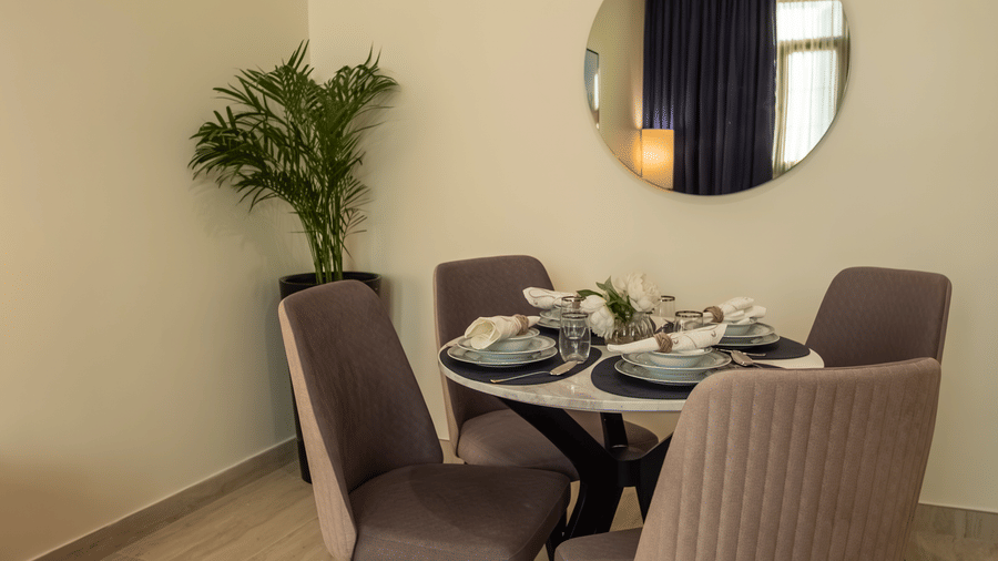 Dining nook with a round white table, four grey upholstered chairs, a round mirror on the wall, and a potted plant.