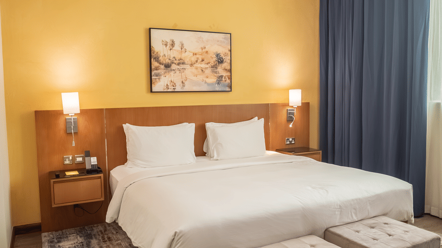 Large bed with white linens, dark wood headboard/nightstands, wall lamps, yellow wall, and two upholstered stools at the foot.