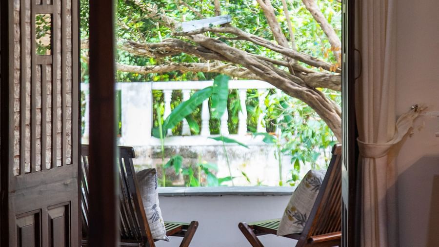 An image of two wooden chairs looking out into greenery with wooden door @ Lamrin Ucassaim Hotel, Goa