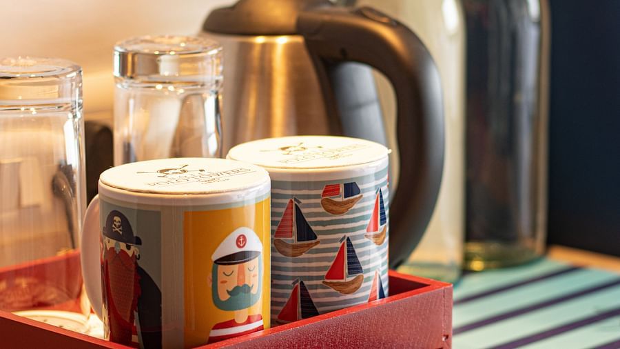 A hotel amenity setup with a kettle, glass bottles, patterned cups, and a selection of snacks on a red tray.
