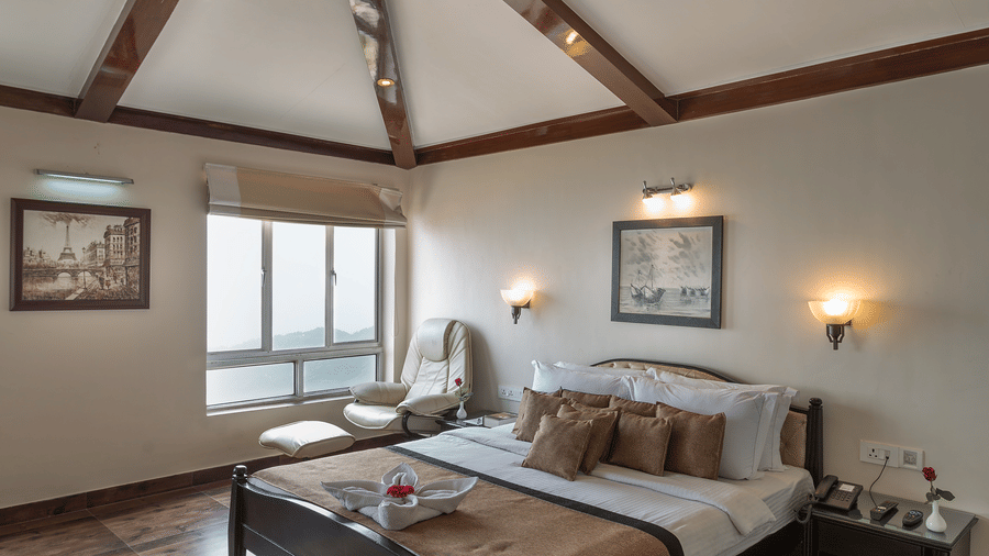 Guest room at Polo Resort Cherrapunji with king bed, exposed beam vaulted ceiling, framed art, shaggy rug, dark hardwood floor and window seat chair.