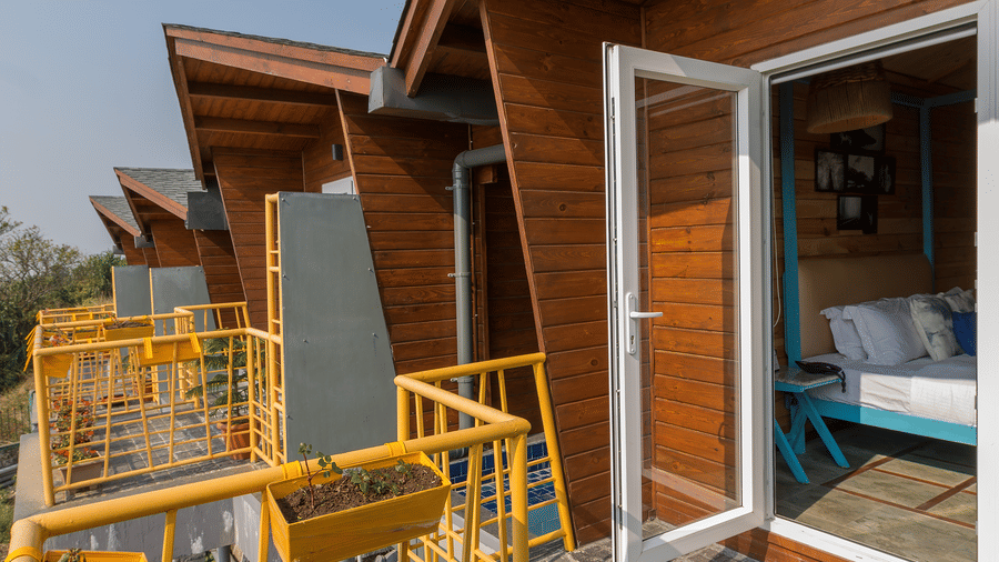 Wooden cottage exterior at Polo Resort Cherrapunji with row of pitched-roof cabins, yellow railings, planter boxes and open door revealing a bed inside.