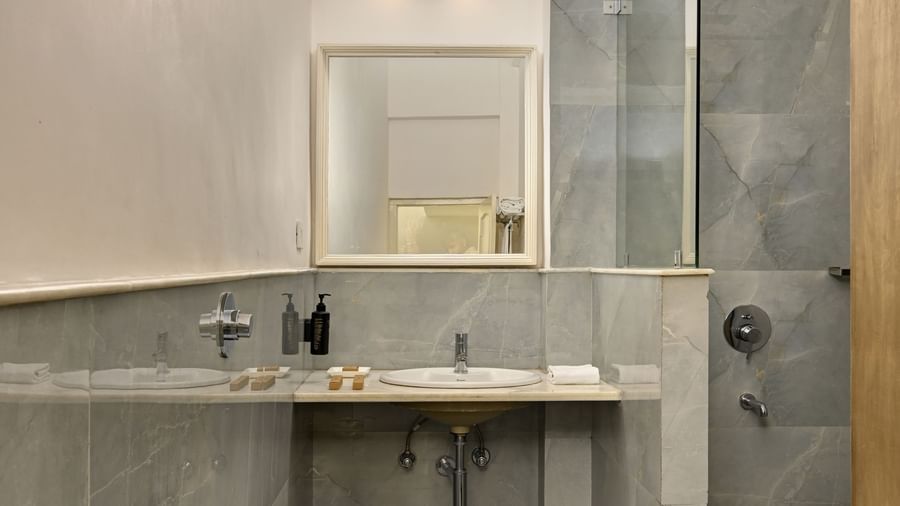 A clean bathroom at Ram Pratap Palace in Udaipur, featuring grey tiled walls and flooring, a western toilet, a countertop with a sink and basic toiletries, a mirror, and a glass-enclosed shower area.
