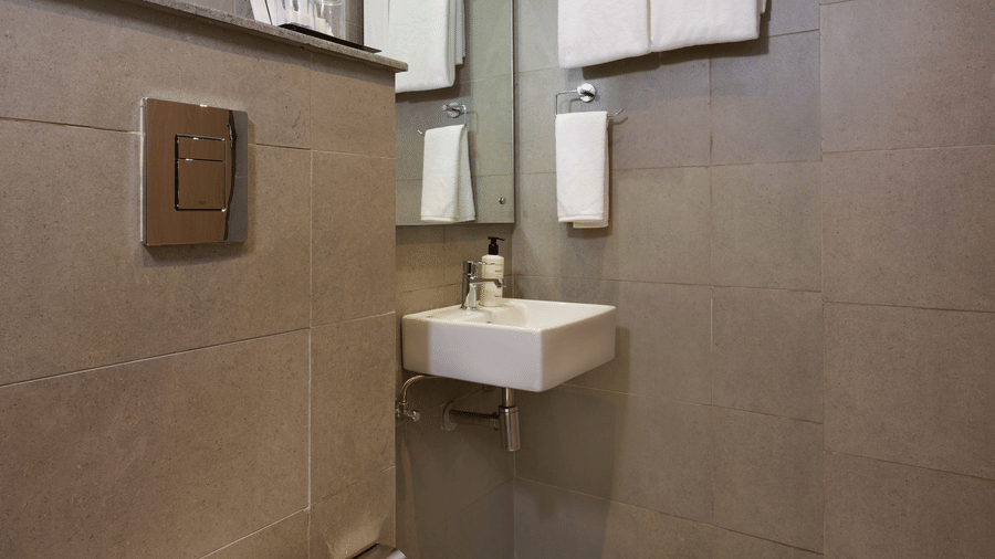 Compact bathroom at Tulip Inn Gurugram with grey tiled walls, a wall-hung commode, a small sink, and a rectangular mirror.