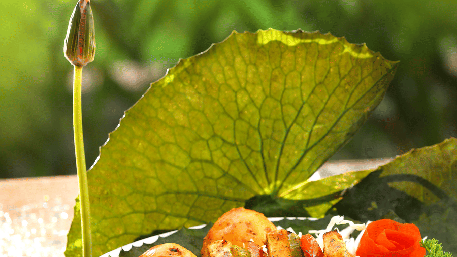 A vegetarian platter sits next to a lotus leaf and bud, while a seafood dish is displayed on the table, with 'Aambal' illuminated on the wall in the background at Uday Backwater Resort