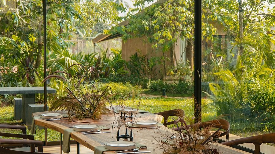 A long, wooden outdoor dining table set with plates and chairs at Villa Moira by V Escapes, under a shaded canopy, surrounded by lush greenery.