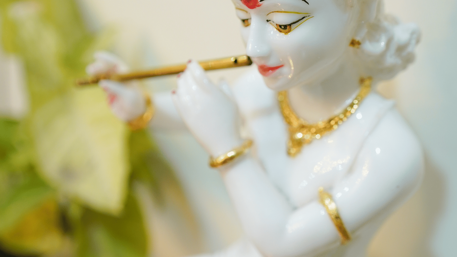 White marble Krishna idol playing a flute, with gold-painted jewellery, beside a green potted plant