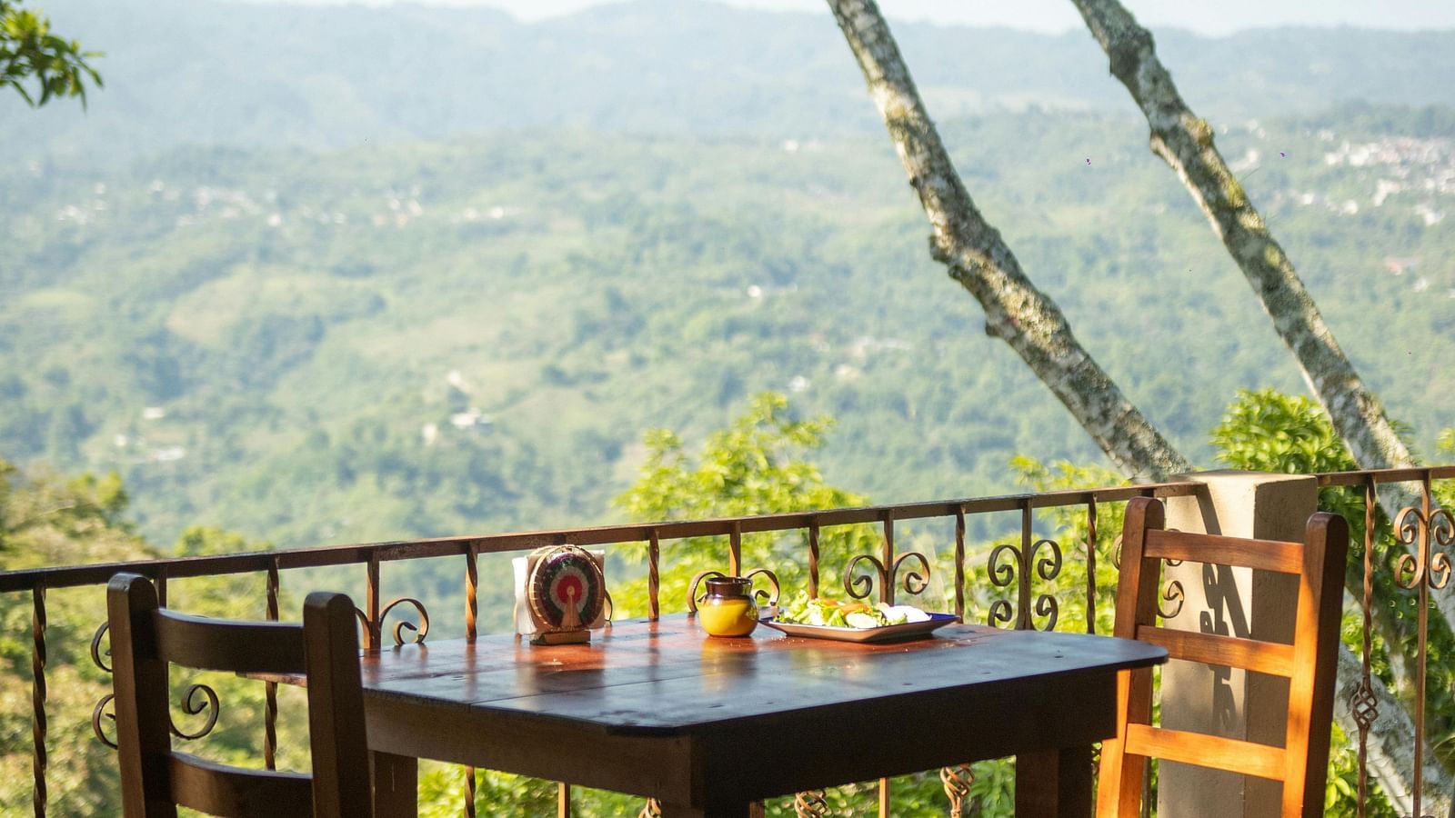 A rustic wooden table and chairs sit on a balcony overlooking a vast, hazy green mountain landscape under a bright sky.