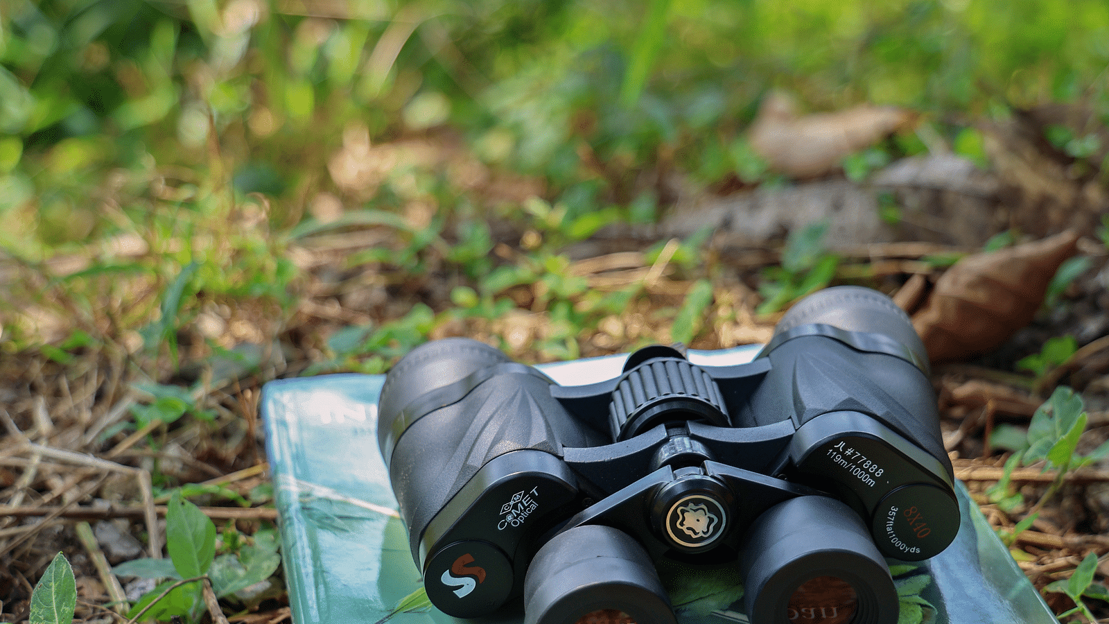 A pair of binoculars placed on a book on grass at The Sylvan Retreat, Dehradun.=