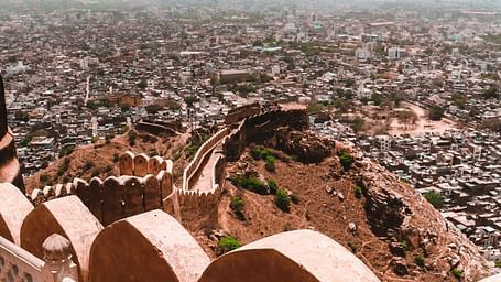 A high-angle view from Nahargarh Fort, Jaipur, overlooking the sprawling city with its densely packed buildings and a distant mountain range under a cloudy sky.