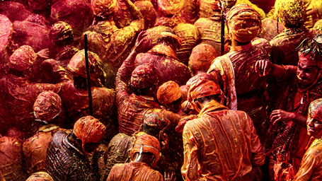 A large crowd celebrates Holi outdoors while drenched in vibrant red and yellow coloured powders.
