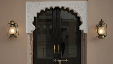 A grand bedroom in the Terrace Suite at Mahendra Niwas, featuring a large bed with a decorative headboard and white bedding. A chandelier hangs from the ceiling.