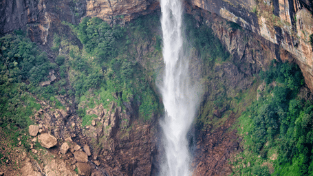 Aerial view of a tall single-drop waterfall plunging from a forested cliff into a turquoise plunge pool surrounded by steep rocky slopes and trees.