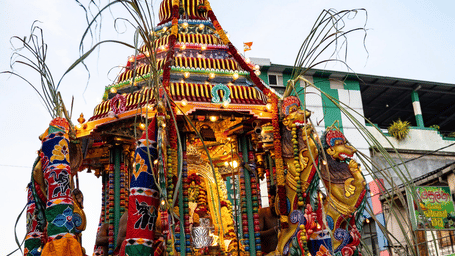 Illuminated and garlanded temple chariot with carved horse figures being pulled through a crowded street during a religious procession.