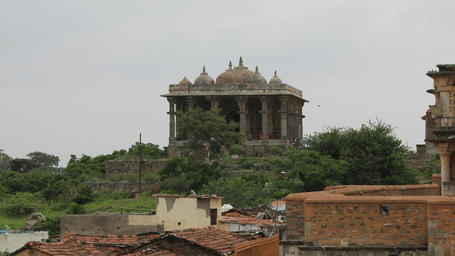An ancient stone temple with multiple domes sits atop a lush green hill, overlooking a small village and pathway