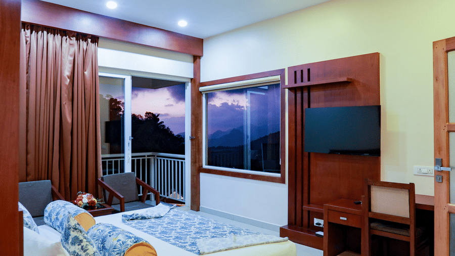 A double bed placed right opposite to a wall-mounted television beside a window framing the mountain views at Aveda Mountains and Mist Resort and Spa, Munnar.
