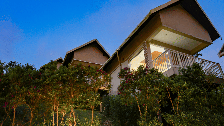 Facade of the Luxe Mountain View Cottage at Aveda Mountains and Mist Resort and Spa, Munnar, with exterior lights lining the green bushes illuminating the outside area during dusk.