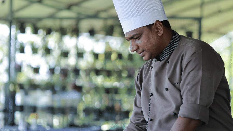 A person wearing an appron and a chef's hat serving the dishes on the table of the Machaan restaurant at Aveda Mountains and Mist Resort and Spa, Munnar.