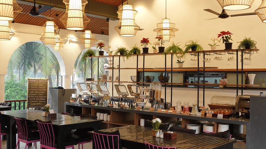 Dining tables, colourful chairs, and a long buffet counter fill a restaurant with arched windows at Bougainvillea, Alibaug.