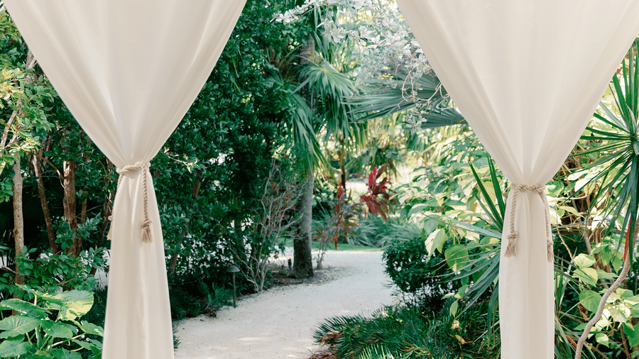 A view through sheer white curtains tied back to reveal a stone garden path winding through lush tropical greenery.