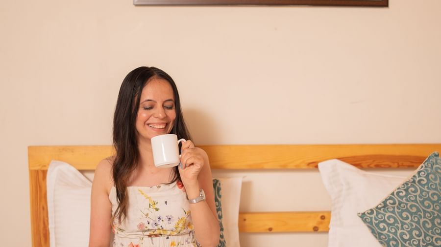 A woman in pyjamas sits on a large bed with a cup, framed by a wooden headboard and a wall art, at Daksh Resort & Amusement Park, Sasan Gir.