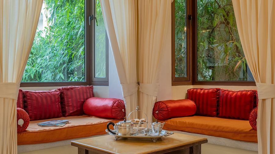 A room with window seats, orange cushions, cream curtains, and a small wooden table on a patterned floor | Dera Rawatsar, Jaipur