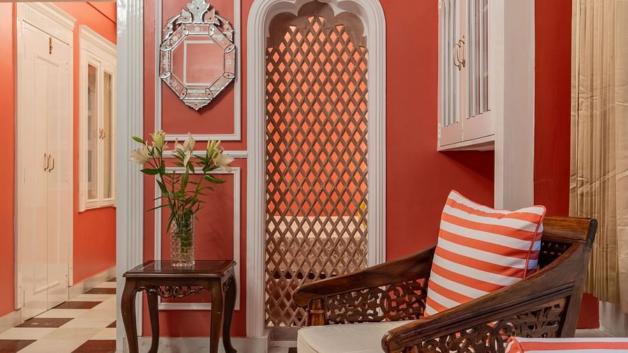 A hallway with red walls, white trim, a checkered floor, a wooden chair with a striped cushion, and an arched doorway | Dera Rawatsar, Jaipur