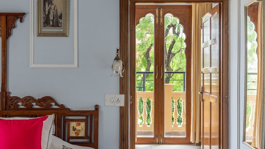 A serene bedroom at Dera Rawatsar, Jaipur, features a comfortable bed, a patterned rug, and an ornate wooden doorway leading to a bright space.