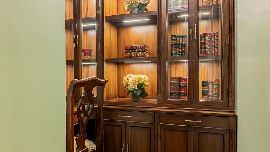 A cozy nook at Dera Rawatsar, Jaipur, features a well-lit, ornate wooden bookcase filled with books and decorative items, beside a wooden chair.