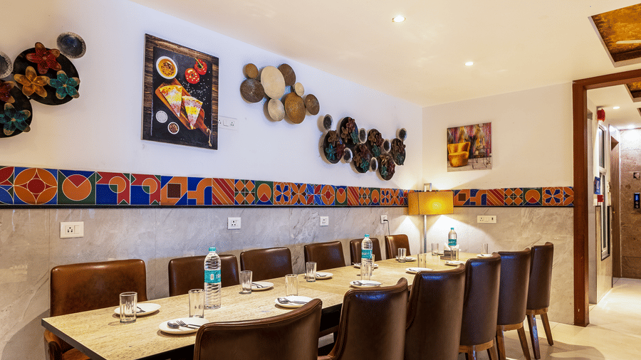 A warm, ambient light illuminating The Curry House, featuring a long dining arrangement with brown leather chairs and decorative wall art at Hotel Corporate Suites