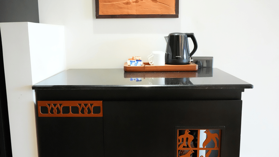 A room area featuring a black cabinet with a kettle on top and a square terracotta wall artwork at Hotel Sonar Bangla, Ajodhya Hills.