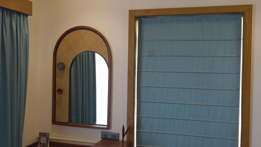  A wooden dressing table with a mirror and a chair set against a white wall with a blue window blind at Hotel Sonar Bangla, Mayapur.