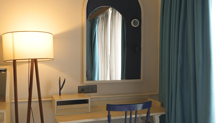 A white writing desk with a blue chair, a floor lamp, and a wall mirror at Hotel Sonar Bangla, Mayapur.