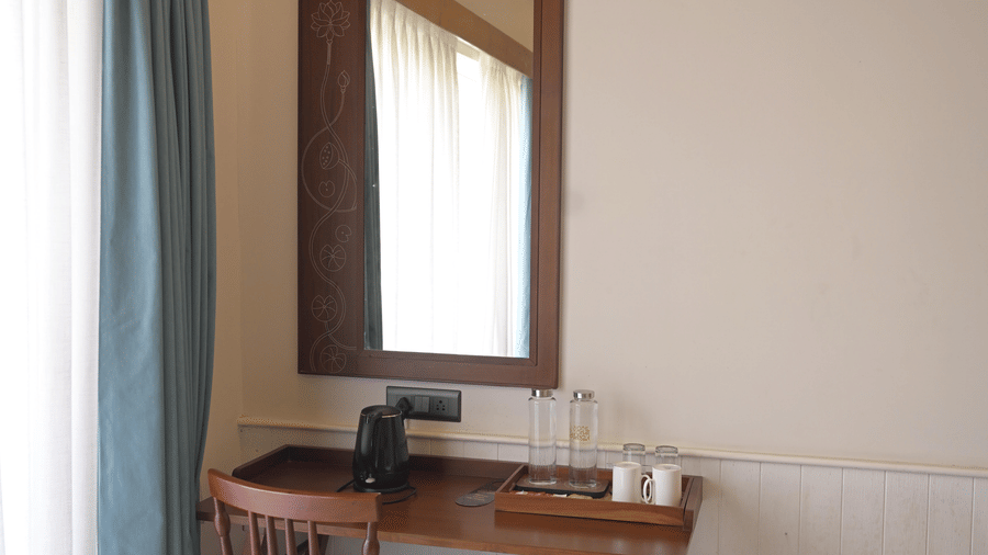 A wooden writing desk with a matching chair and a wall-mounted mirror next to blue curtains at Hotel Sonar Bangla, Mayapur. 
