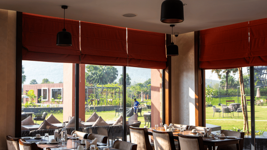 Sunlit corner dining area at Tattva Restaurant with wooden tables, cream upholstered chairs, pendant lights, and large windows overlooking the green lawn | Hotel Sonar Bangla, Susunia