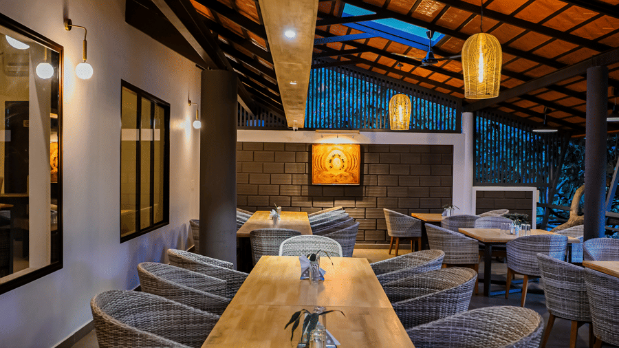 A long light wooden dining table set with grey woven chairs under a high sloped ceiling with exposed wooden rafters at Kadkani River Resort, Coorg.