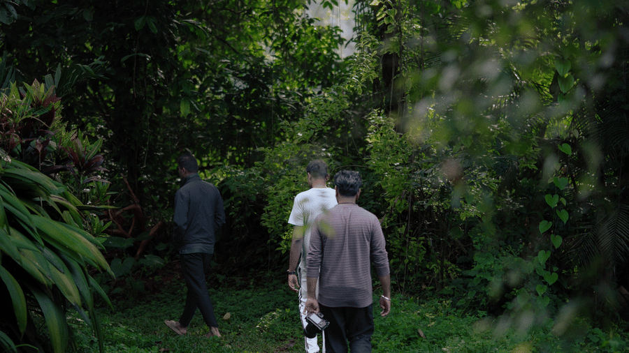 3 people walking along a narrow grass pathway surrounded by dense green trees, bushes, and overhanging foliage at Kadkani River Resort, Coorg.