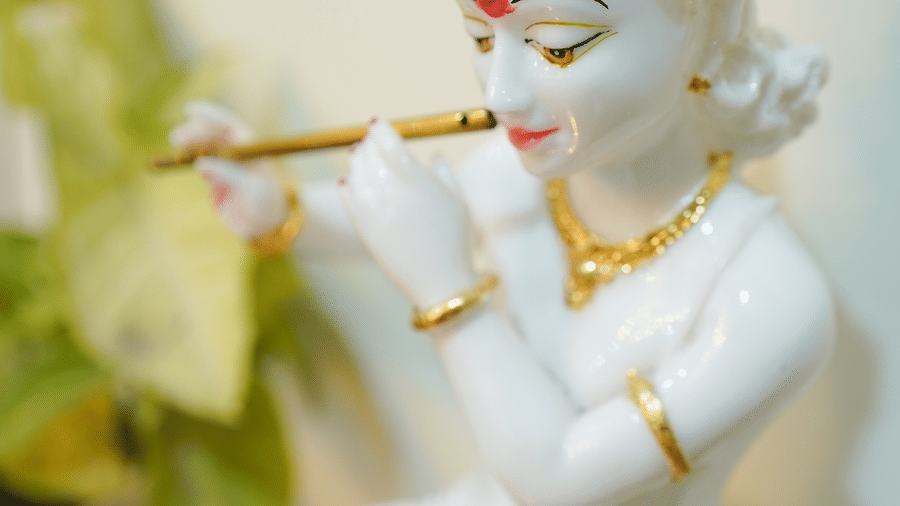 Close-up of a white marble Krishna idol playing a flute, with gold-painted jewellery and a green crown leaf, beside a green potted plant